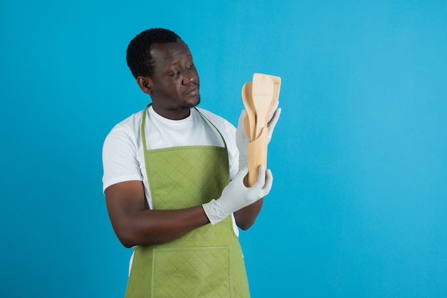 Imagen de un hombre en delantal verde sosteniendo utensilios de cocina de madera contra la pared azul