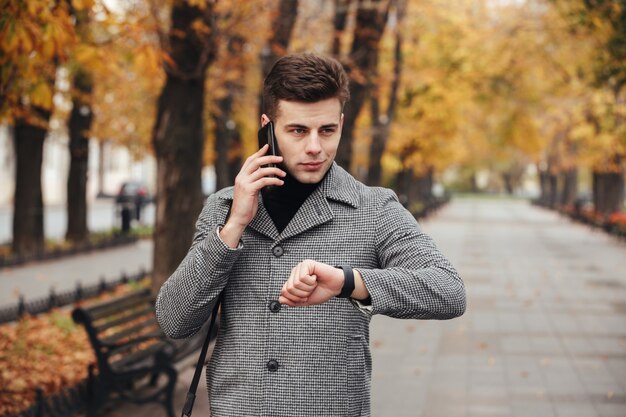 Imagen de hombre elegante que controla el tiempo con reloj en mano y hablando por teléfono móvil durante su caminata en el parque