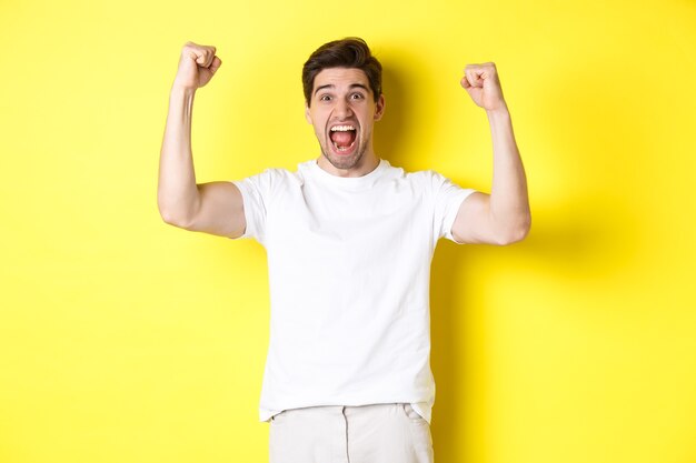 Imagen de hombre emocionado ganando, levantando las manos y celebrando, triunfando y animando al equipo, de pie sobre fondo amarillo. Copia espacio