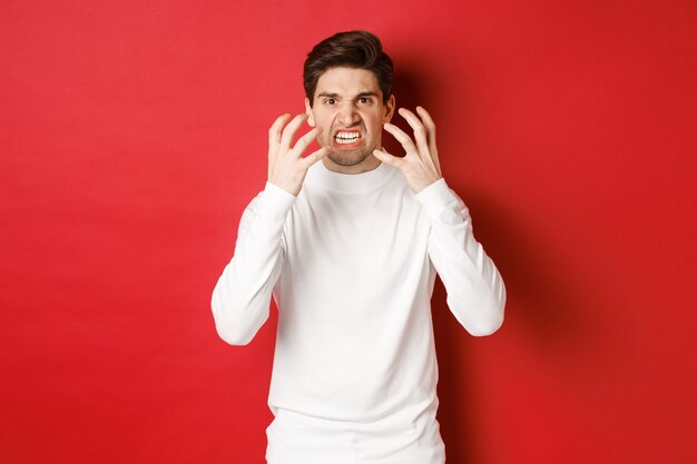 Imagen de hombre enojado y cabreado en suéter blanco haciendo muecas y temblando de rabia de pie furioso ...