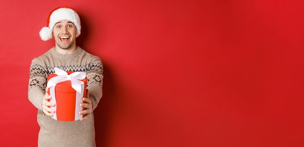 Imagen de hombre feliz diciendo feliz Navidad y dándote un regalo en caja, sonriendo alegre, celebrando las vacaciones de invierno con gorro de Papá Noel y suéter de Navidad, de pie sobre fondo rojo.