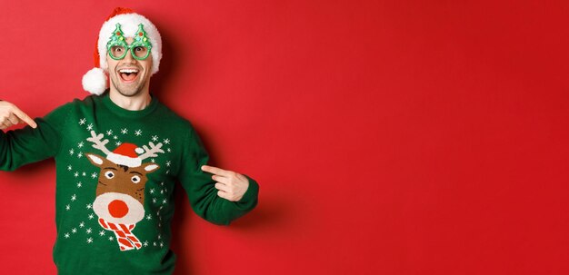 Imagen de un hombre feliz con gafas de fiesta y gorro de Papá Noel señalando su suéter navideño y sonriendo de pie sobre un fondo rojo