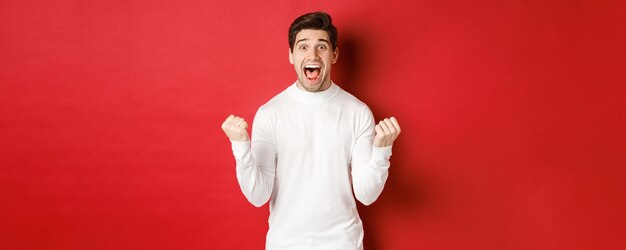 Imagen de un hombre feliz y guapo con suéter blanco, ganando algo, haciendo puñetazos y sonriendo asombrado, celebrando la victoria, de pie sobre un fondo rojo.