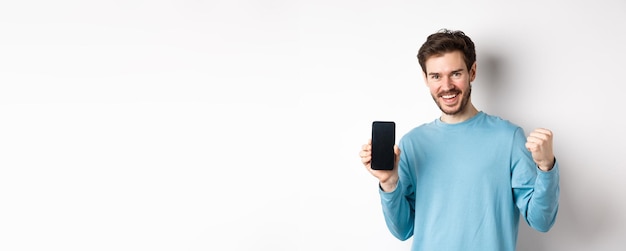 Foto gratuita imagen de un hombre feliz que muestra la pantalla vacía del teléfono inteligente y celebra sonriendo con regocijo y puño pum