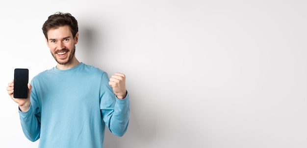 Foto gratuita imagen de un hombre feliz que muestra la pantalla vacía del teléfono inteligente y celebra sonriendo con regocijo y puño pum