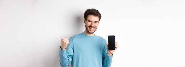 Foto gratuita imagen de un hombre feliz que muestra la pantalla vacía del teléfono inteligente y celebra sonriendo con regocijo y puño pum