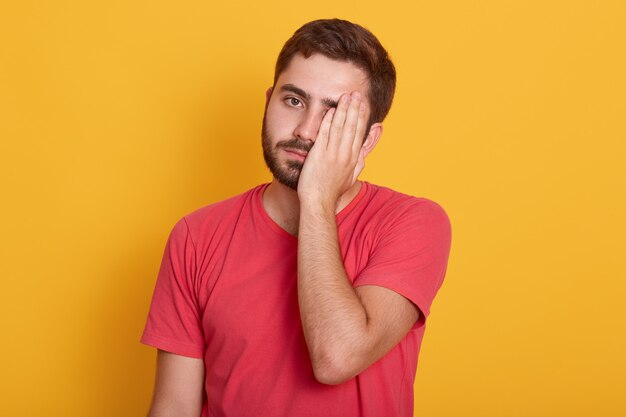 Imagen de hombre guapo con camiseta roja informal, de pie con expresión triste, cubriendo la mitad de su rostro con la mano, parece cansado