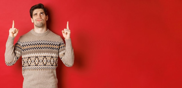 Imagen de un hombre guapo decepcionado y escéptico, vestido con un suéter de Navidad, sonriendo y frunciendo el ceño mientras mira algo malo, señalando con el dedo hacia arriba, de pie sobre un fondo rojo