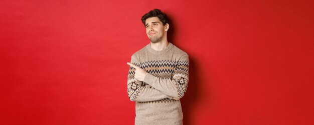 Imagen de un hombre guapo decepcionado con suéter navideño, mirando y señalando a la izquierda con la cara molesta, expresa arrepentimiento, mostrando un anuncio de año nuevo, de pie sobre un fondo rojo