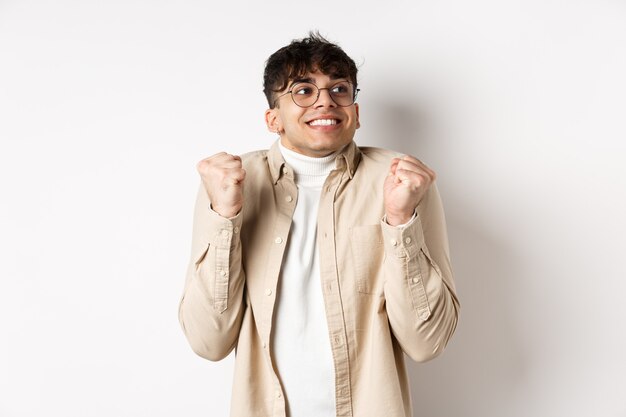 Imagen de hombre guapo emocionado que se siente motivado y afortunado, mirando a la derecha y sonriendo, haciendo un gesto de bomba de puño para celebrar la victoria, ganando el premio, de pie sobre fondo blanco.