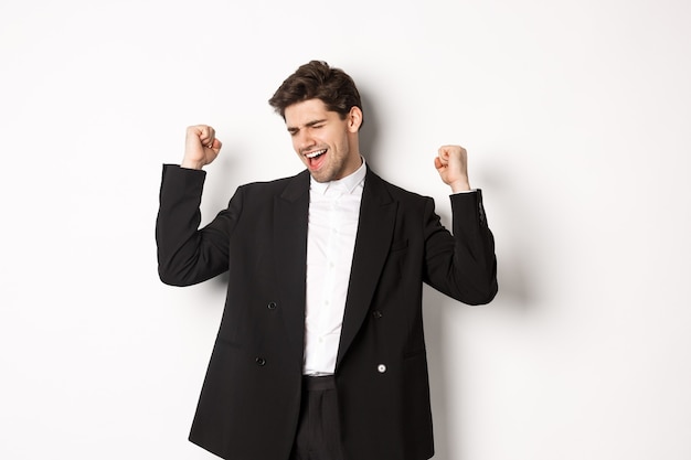 Imagen de hombre guapo exitoso y complacido en traje, regocijándose y haciendo señales de bomba de puño, bailando de felicidad, de pie sobre fondo blanco.