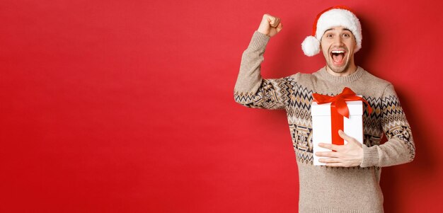 Imagen de un hombre guapo feliz y emocionado recibiendo un regalo de navidad levantando las manos triunfante y sonriendo celebrando el año nuevo de pie sobre un fondo rojo con sombrero de santa