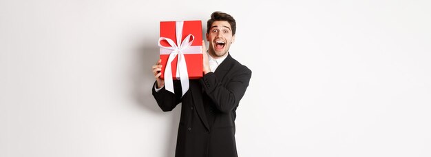 Imagen de un hombre guapo feliz con traje negro sosteniendo una caja con un regalo de Navidad y sonriendo de pie