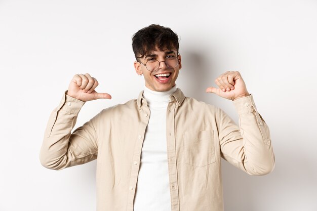 Imagen de un hombre guapo sonriente motivado apuntando a sí mismo, autopromoción y con apariencia de confianza, de pie sobre fondo blanco.