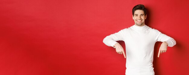 Imagen de un hombre guapo sonriente con suéter blanco invitando a visitar la página, señalando con el dedo hacia abajo y mostrando la pancarta navideña, parado en un fondo rojo.