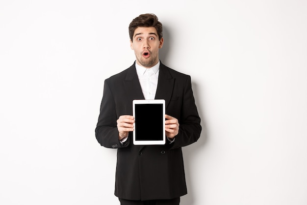 Imagen de un hombre guapo sorprendido en traje negro, mostrando la pantalla de la tableta digital y mirando asombrado, de pie contra el fondo blanco.