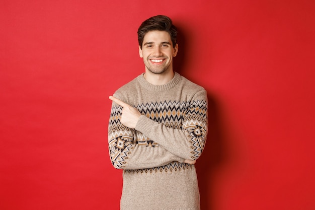 Imagen de un hombre guapo en suéter navideño, celebrando las vacaciones de año nuevo, sonriendo feliz y señalando con el dedo en el espacio de copia de la esquina superior izquierda, de pie sobre fondo rojo