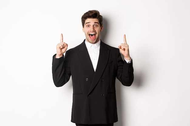 Imagen de un hombre guapo en traje de fiesta, mostrando la promoción de vacaciones, apuntando con el dedo hacia arriba y sonriendo asombrado, de pie sobre fondo blanco.