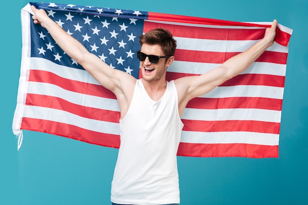 Imagen del hombre hermoso joven que se coloca sobre azul aislado sosteniendo la bandera de los EEUU Mirando a un lado
