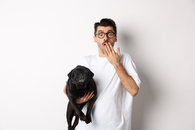 Imagen de hombre joven guapo con perro lindo y jadeando sorprendido. Dueño de la mascota mirando a la cámara conmocionado, lleva pug negro en el brazo, fondo blanco.