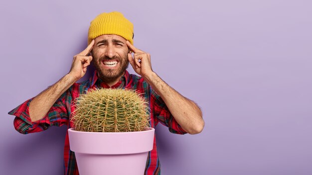 La imagen de un hombre molesto mantiene ambos dedos índices en las sienes, sufre de migraña, sonríe de dolor, trata de concentrarse, crece cactus en macetas, posa sobre un fondo morado.