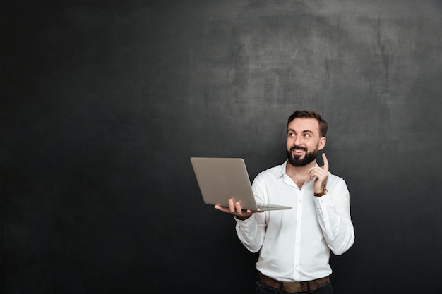 Imagen de hombre morena inteligente tiene idea gesticulando con el dedo hacia arriba mientras usa una computadora portátil plateada, aislada sobre una pared gris oscuro