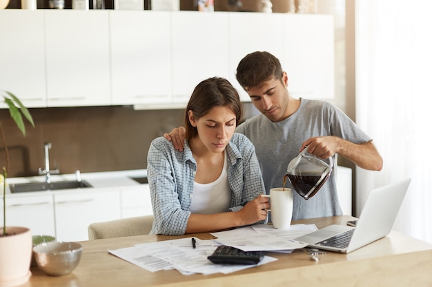 Imagen de un hombre y una mujer jóvenes haciendo papeleo juntos en casa: esposa seria sentada en la mesa de comedor con papeles y computadora portátil, calculando las facturas mientras su esposo le sirve café