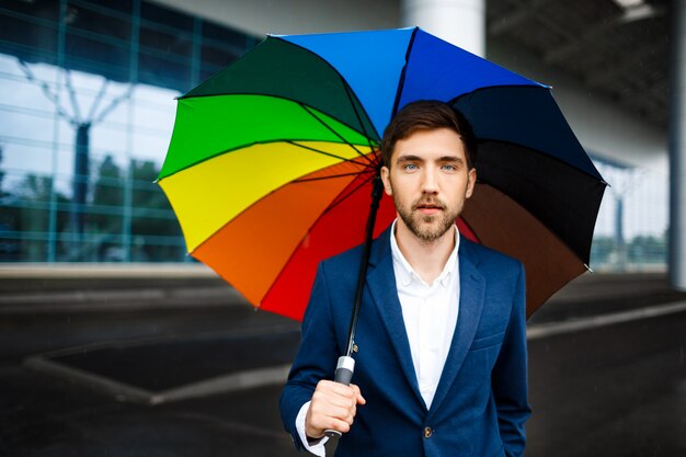 Imagen del hombre de negocios joven confiado que sostiene el paraguas abigarrado en la calle