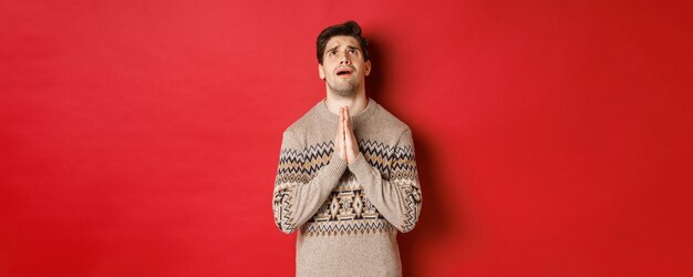 Imagen de un hombre nervioso y esperanzado rezando a Dios, rogando ayuda con la Navidad, usando suéter de invierno, mirando hacia arriba y suplicando, de pie sobre un fondo rojo