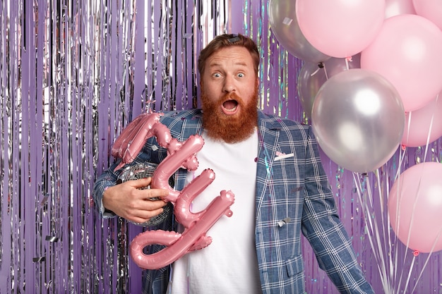 Imagen de un hombre pelirrojo asustado que lleva una bola de discoteca brillante, globos, celebra la promoción en un club nocturno con sus colegas, sorprendido con mala música, viste un elegante abrigo a cuadros, aislado en una pared púrpura