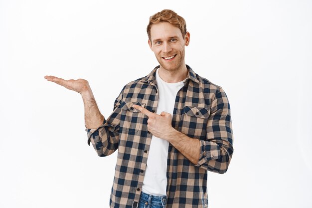 Imagen de un hombre pelirrojo sonriente apuntando a su mano abierta, mostrando un artículo, recomendando un producto en su palma, mostrando el objeto, de pie contra la pared blanca