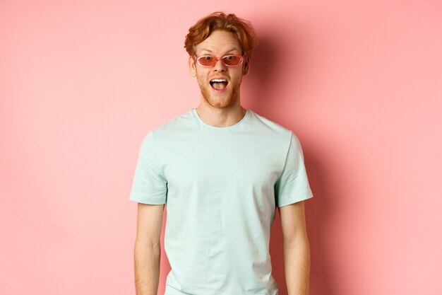 Imagen de hombre pelirrojo sorprendido de vacaciones, con gafas de sol con camiseta de verano, boca abierta y diciendo wow asombrado, de pie sobre fondo rosa.