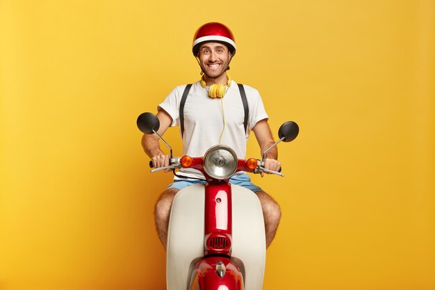 Imagen de hombre positivo monta scooter, usa casco, camiseta blanca, está de buen humor, aislado sobre la pared amarilla del estudio