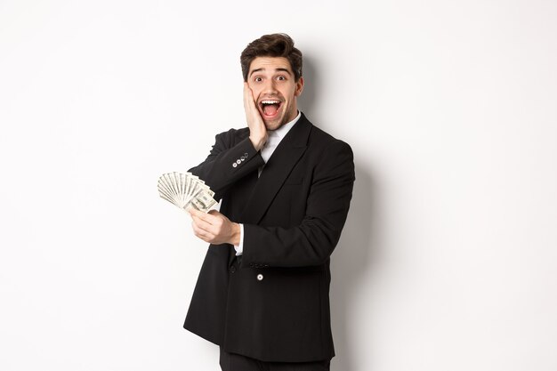 Imagen de hombre rico y feliz en traje negro, ganador del premio, sosteniendo dinero y mirando emocionado a la cámara, de pie sobre fondo blanco.