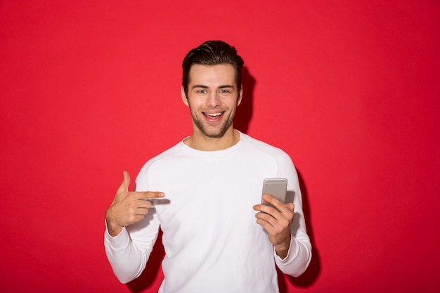 Imagen del hombre sonriente en suéter mirando mientras sostiene el teléfono inteligente y apuntando a él sobre la pared roja
