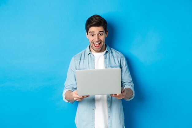 Imagen de hombre sorprendido y feliz reaccionando a una oferta especial en internet, mirando el portátil emocionado, de pie contra el fondo azul.