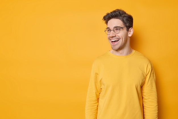 Imagen horizontal de un apuesto hombre moreno que usa anteojos redondos, un saltador casual se ve feliz y ve algo divertido aislado sobre un fondo amarillo vívido con espacio en blanco para su promoción