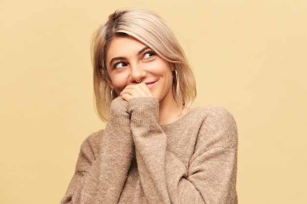 Imagen horizontal de atractiva encantadora joven mujer caucásica con anillo en la nariz, aretes redondos y elegante suéter beige cálido tomados de la mano y mirando a otro lado con una sonrisa misteriosa