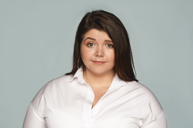 Foto gratuita imagen horizontal de una atractiva joven con sobrepeso de talla grande vestida con camisa blanca formal y aretes redondos posando en la pared gris en blanco, con expresión facial preocupada, desconcertada o frustrada