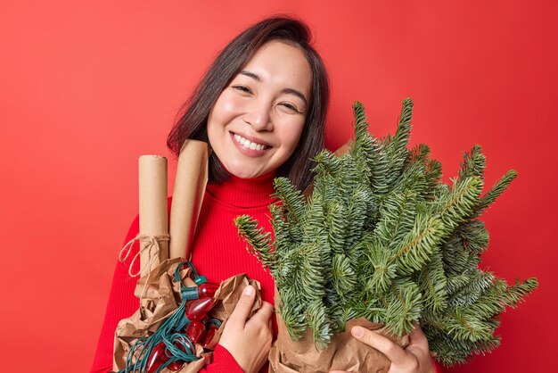 Imagen horizontal de una guapa morena asiática con una sonrisa suave en la cara que inclina la cabeza posa con atributos de vacaciones que se prepara para el Año Nuevo aislado sobre un fondo rojo vívido. se acerca la navidad