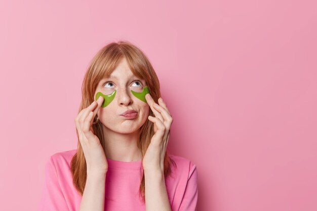 Imagen horizontal de una hermosa pelirroja europea que se pone parches de hidrogel verde debajo de los ojos enfocados sobre vestidos casualmente posa contra un espacio de copia en blanco de fondo rosa para su información