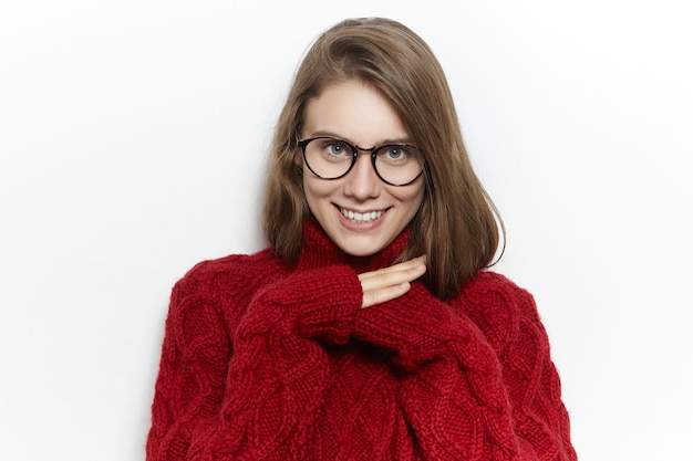 Imagen horizontal de una linda adolescente alegre con un suéter de cuello alto granate mientras pasa el frío día de otoño en casa, calentando, escondiendo las manos dentro de las mangas y sonriendo felizmente