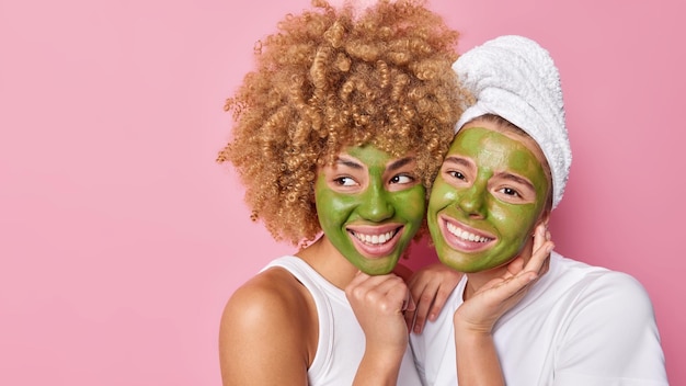 Imagen horizontal de mujeres bastante jóvenes con alegres expresiones de ensueño aplicar máscaras naturales verdes para el tratamiento de la piel usar camisetas blancas casuales aisladas sobre fondo rosa espacio en blanco