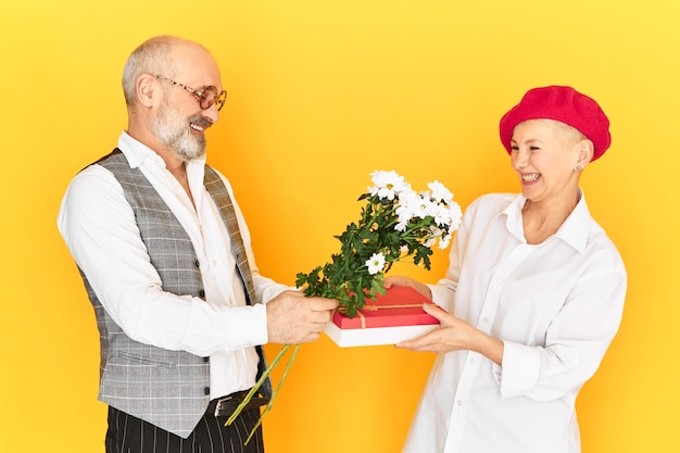 Imagen horizontal del tímido abuelo torpe con barba gris sosteniendo flores y caja de regalo felicitando a su novia madura en su cumpleaños. Linda pareja de ancianos feliz en la primera cita