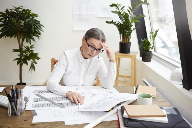 Imagen de una ingeniera de mediana edad estresada y molesta con camisa blanca y gafas mirando planos o documentación del proyecto frente a ella en el escritorio, frustrada al ver tantos errores