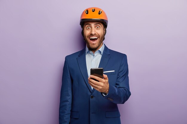 Imagen de ingeniero feliz sostiene teléfono móvil, envía mensajes de texto a colegas, usa casco naranja y traje elegante