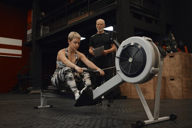 Imagen de un instructor de fitness masculino barbudo senior con portapapeles viendo a su clienta hacer ejercicio en la máquina de remo. Mujer atractiva entrenando en el gimnasio con entrenador personal, haciendo ejercicios de cardio