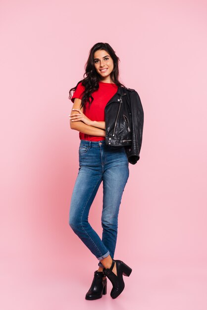 Imagen integral de la sonriente mujer morena posando con los brazos cruzados y mirando a la cámara sobre rosa