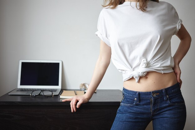 Imagen interior de una chica delgada de moda posando en casa, vestida con jeans y camiseta blanca con un espacio vacío para su diseño, colocando la mano sobre el escritorio con gafas y una computadora portátil con pantalla en blanco