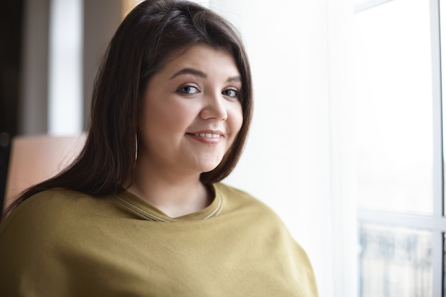 Imagen interior de encantadora elegante joven mujer de veinte años con mejillas regordetas en elegante sudadera y aretes, con aspecto alegre